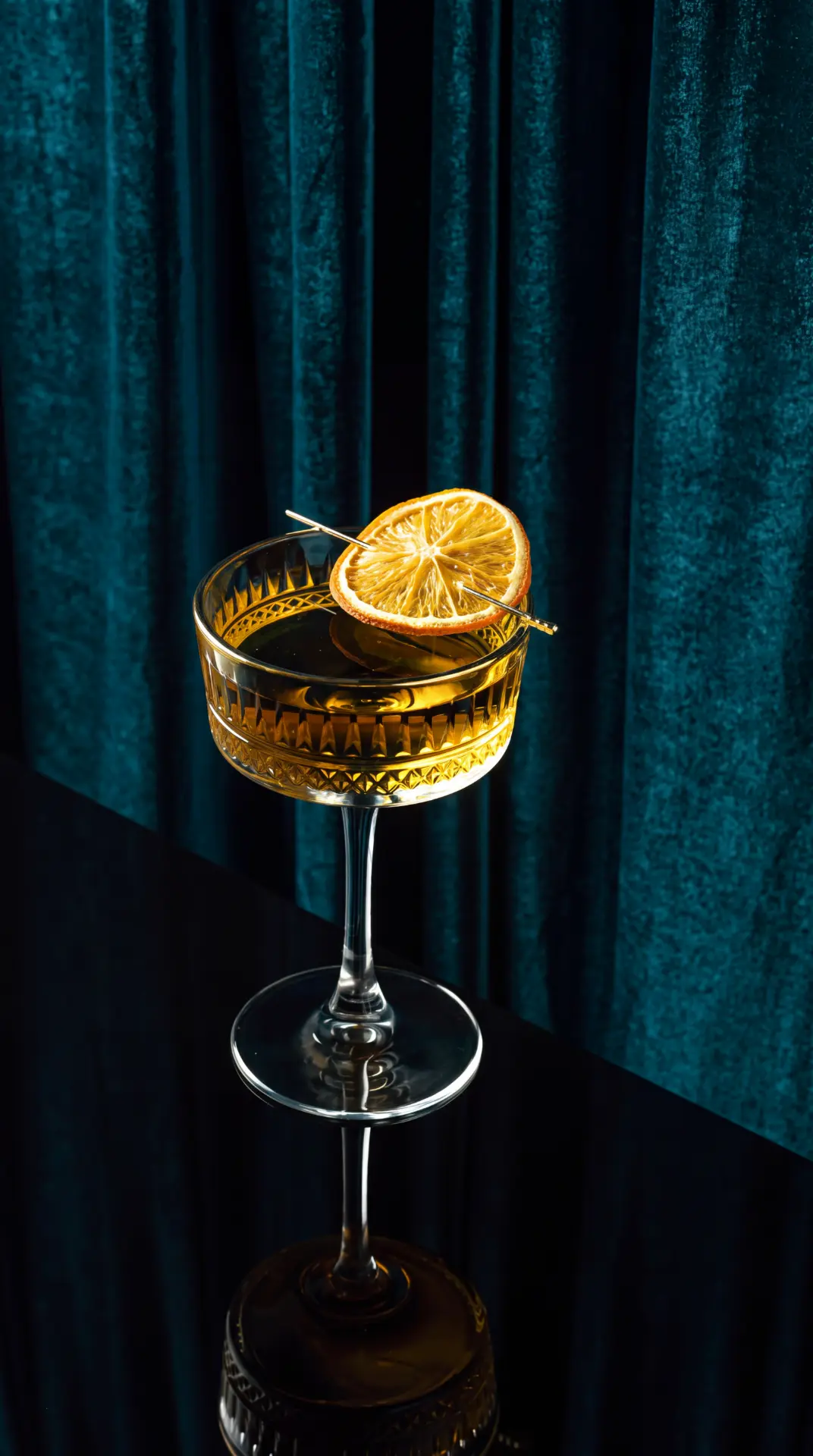 Cocktailglas mit bernsteinfarbenem Drink und getrockneter Orangenscheibe vor Samtvorhang